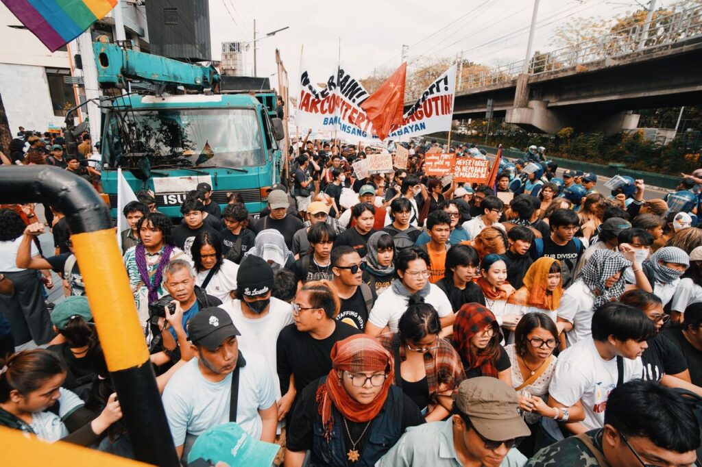 A diverse crowd gathered in an urban street for a protest, displaying banners and flags.